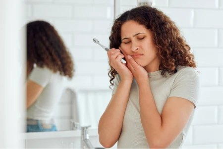 Person standing in a bathroom clutching their cheek in pain while holding a toothbrush, as if suffering from a severe toothache.